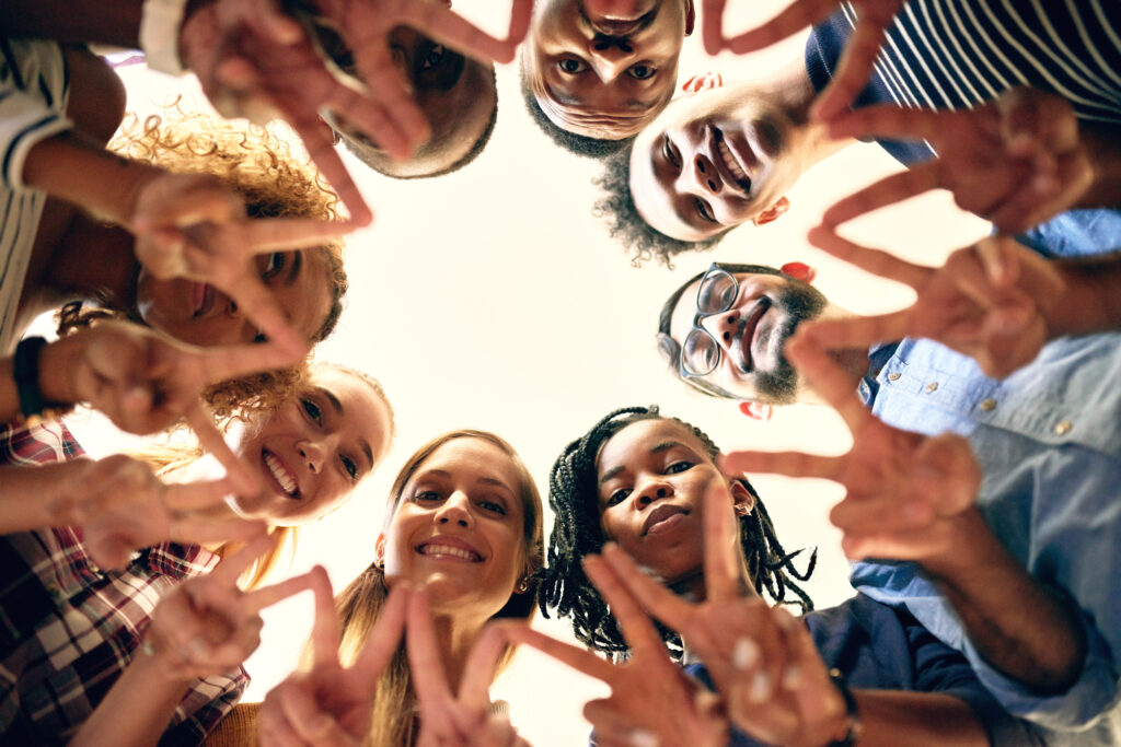 Image of a group of people standing in a circle, looking down at the camera with smiles on their faces, while giving peace signs with their hands. Their gesture symbolizes unity and solidarity in the fight against social stigma surrounding mental health and addiction.