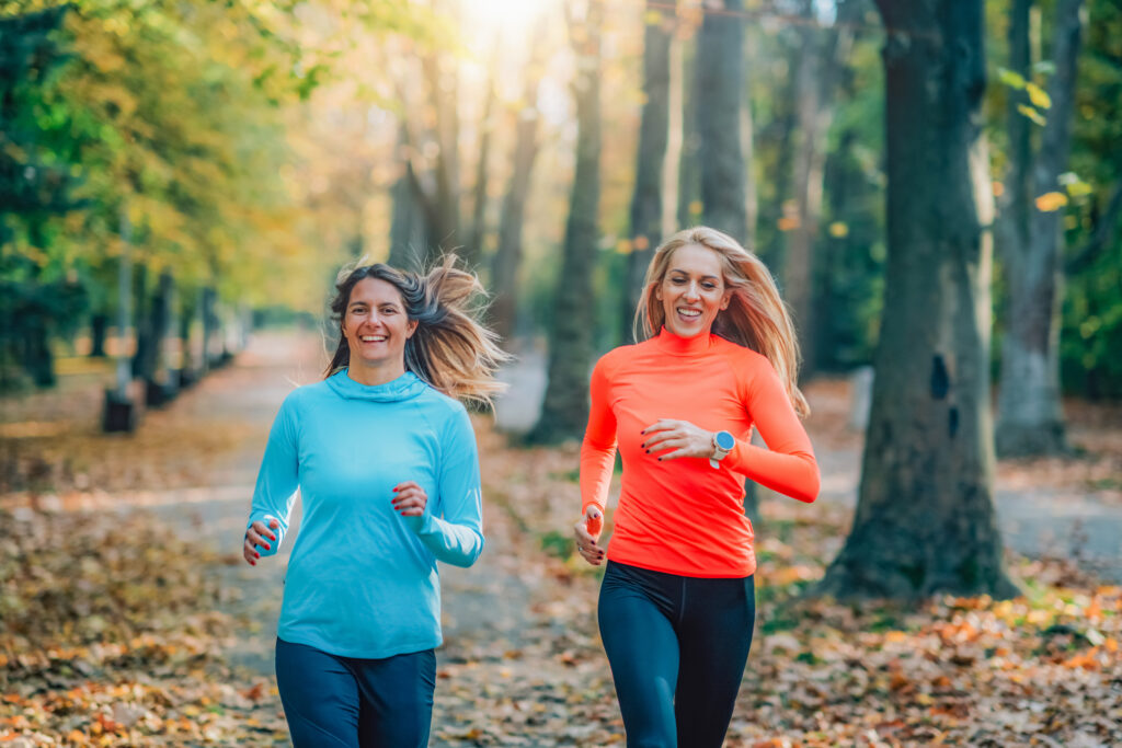 Two smiling women jogging in a park, representing the positive impact of physical activity on mental health.

