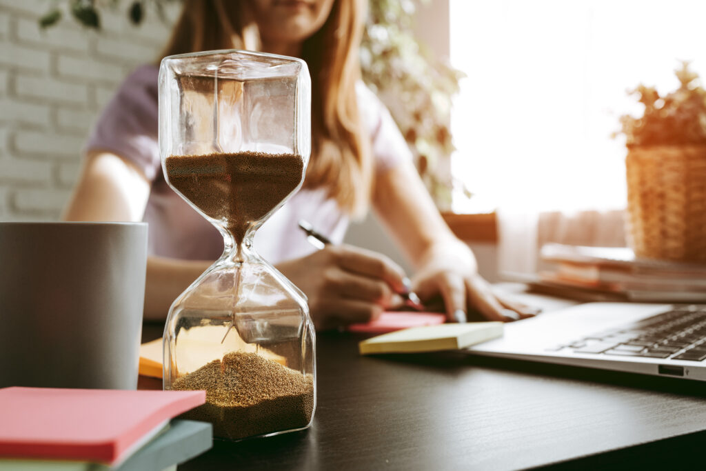 Hourglass on a desk with a woman working in the background, representing daily strategies for adults with ADHD to improve time management.