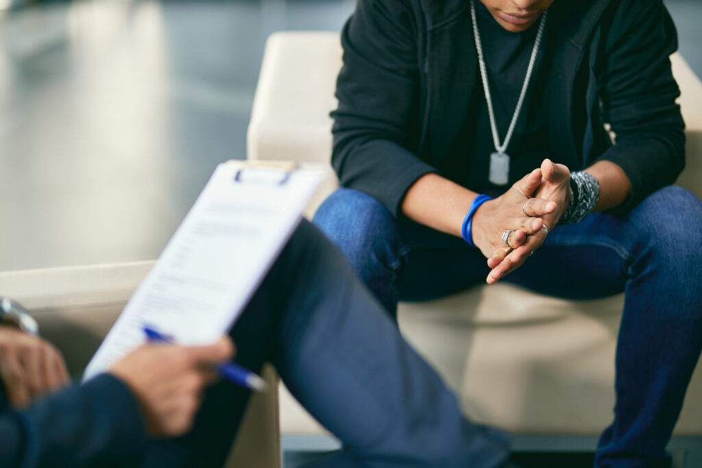 A young person clasps their hands anxiously during a one-on-one therapy session with a counselor.